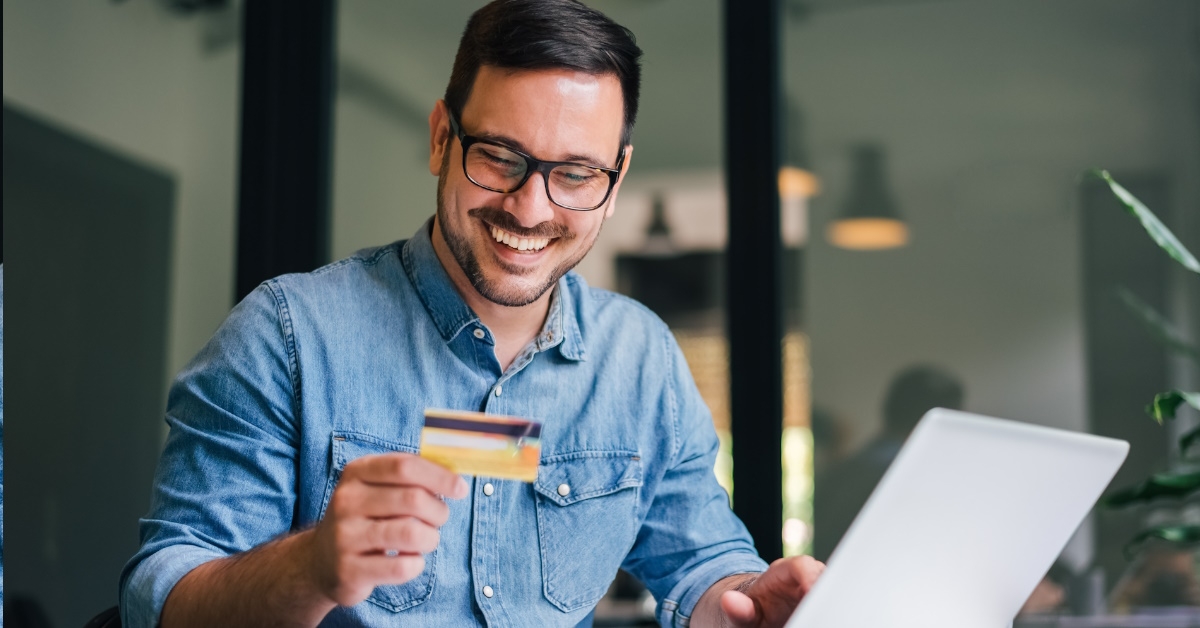 An entrepreneur holding his debit card is making an online payment for goods or services with his laptop on the table and appears to be satisfied.