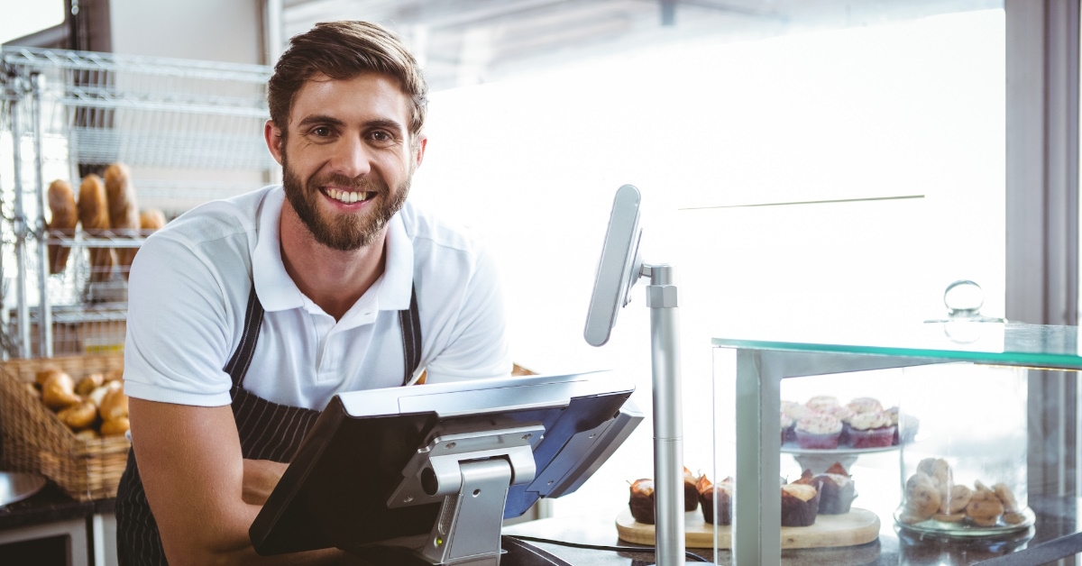 A worker wearing an apron is standing on a bakery counter, posing for the camera with a smile while surrounded by many baked items in this image.