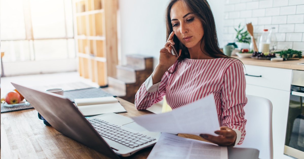 A woman is sitting on a table in her kitchen, talking on her mobile phone and reading a paper document with a thoughtful expression.
