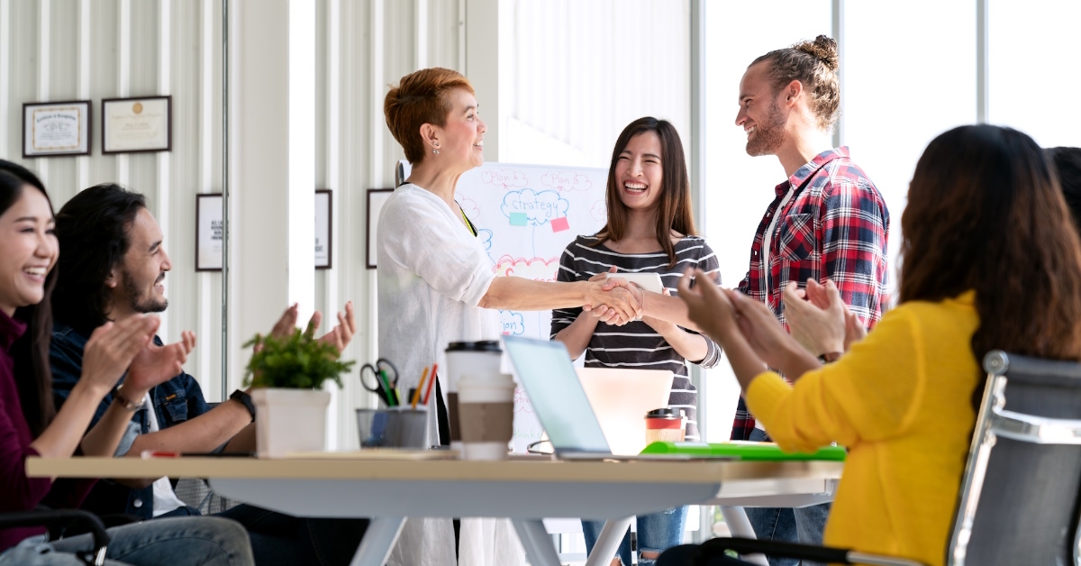 Asian woman shakes hands with a young, bearded, hipster Caucasian man to welcome him as a new employee while the team applauds in support,