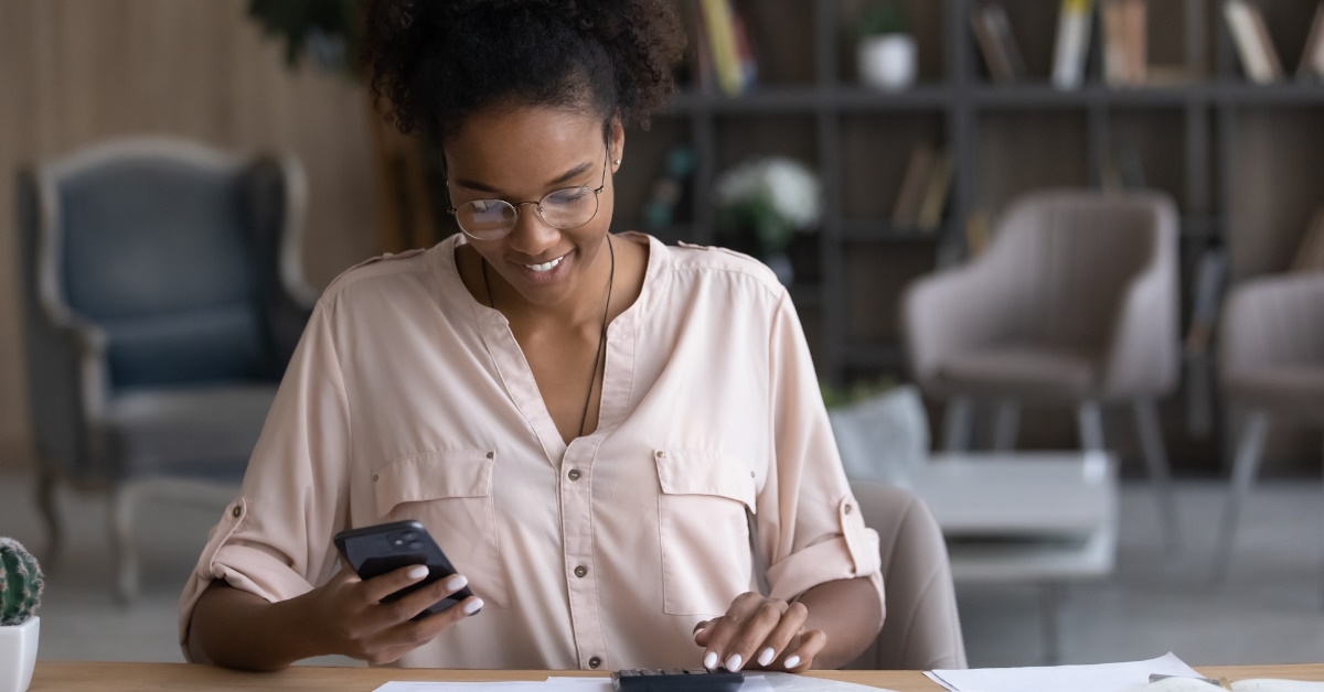  A smiling African American woman wearing glasses is using her smartphone to browse online banking services while calculating bills with a calculator.