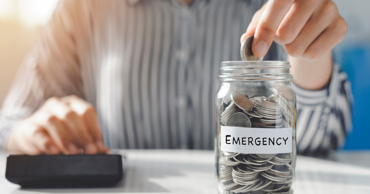 A young woman's hand placing coins in a glass jar on a table to save money for emergency situations.