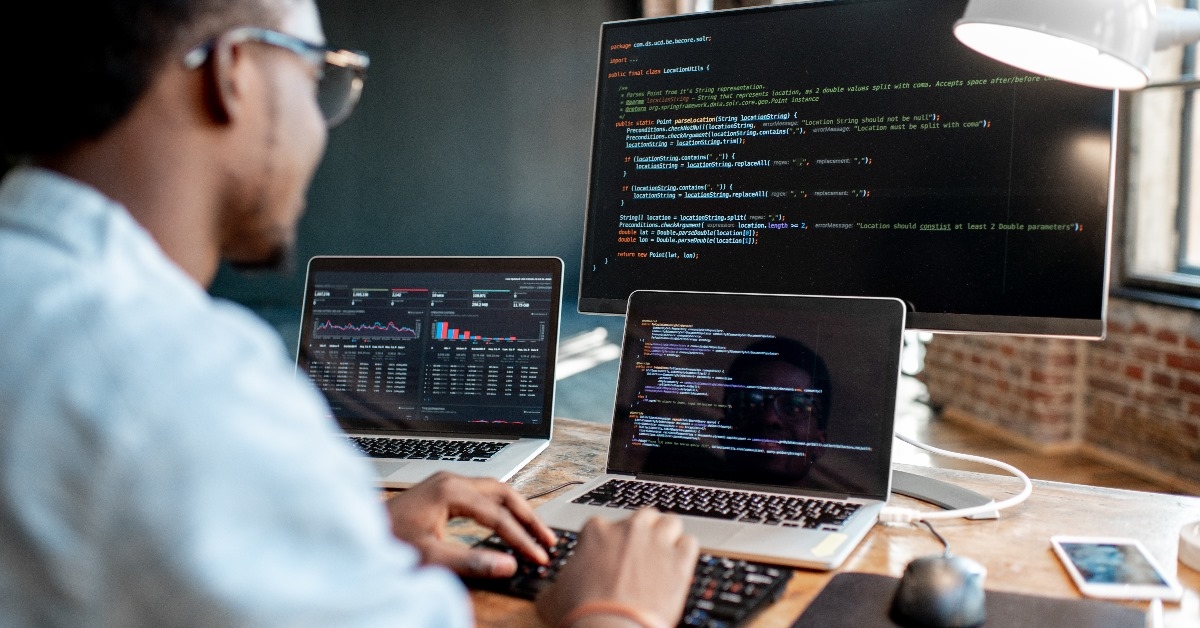 A young African male programmer is running program and observing outputs on three interconnected monitors through an interface while sitting at his workplace in the office.