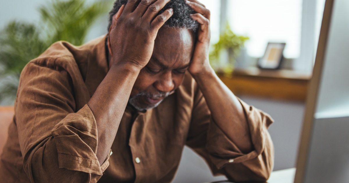 senior african american man sitting at table holding head in stress while looking at notebook