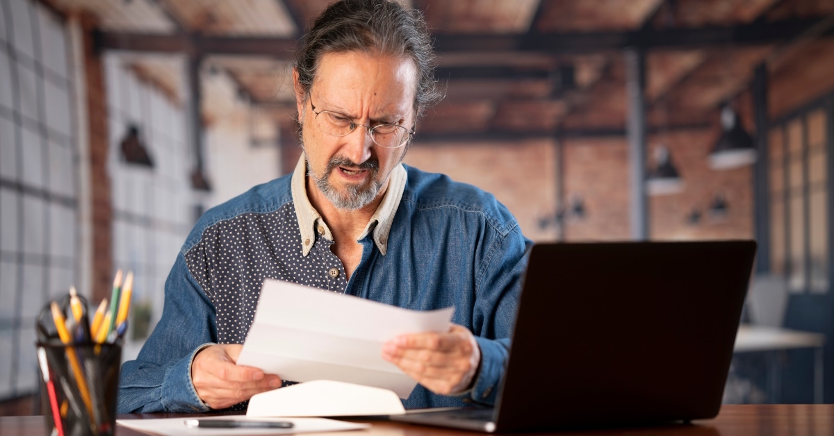 senior man sitting at table in home office reviewing bills while using laptop