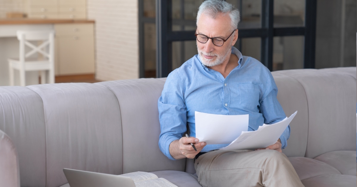 senior man sitting on couch reviewing bills while using laptop