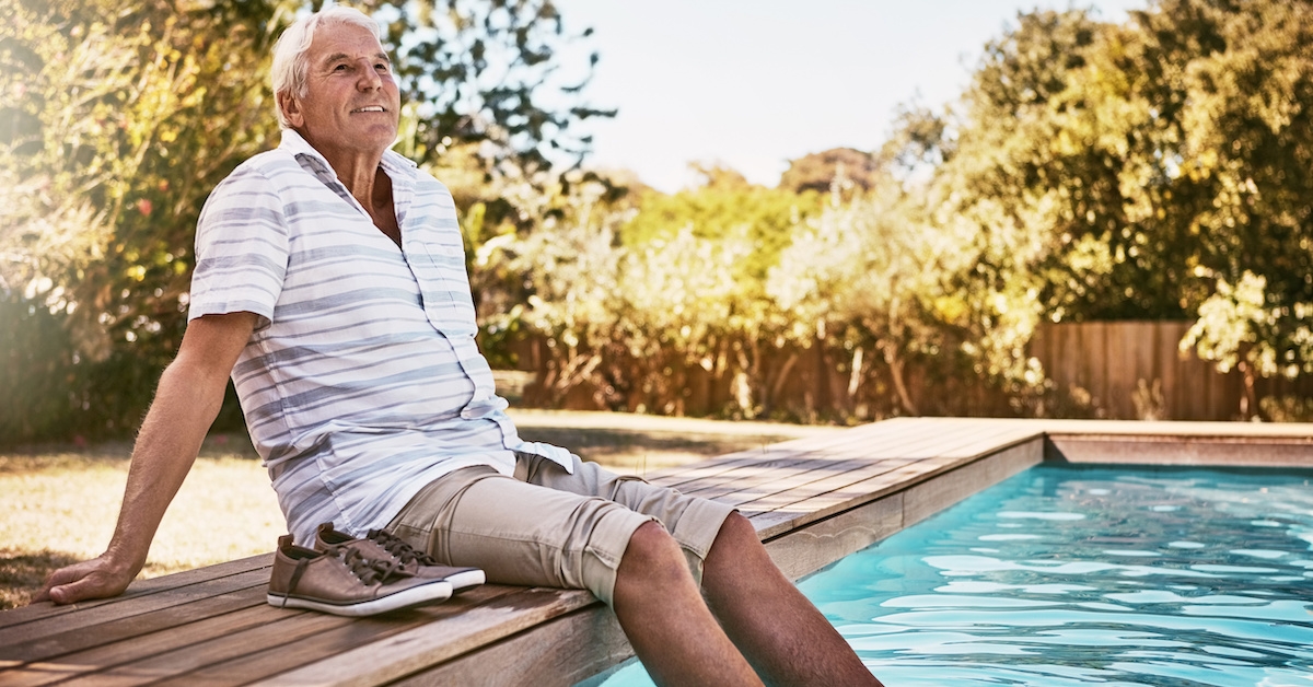 A senior man sits next to the pool and dips his feet in