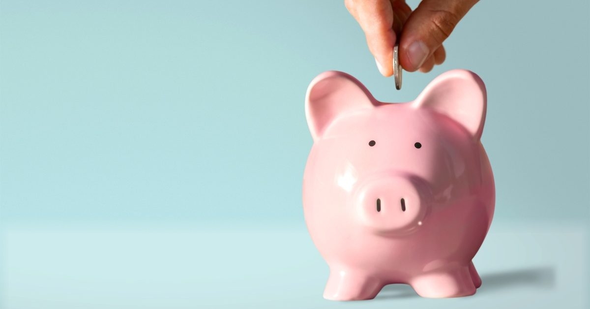 A hand puts a coin into a pink ceramic piggy bank.