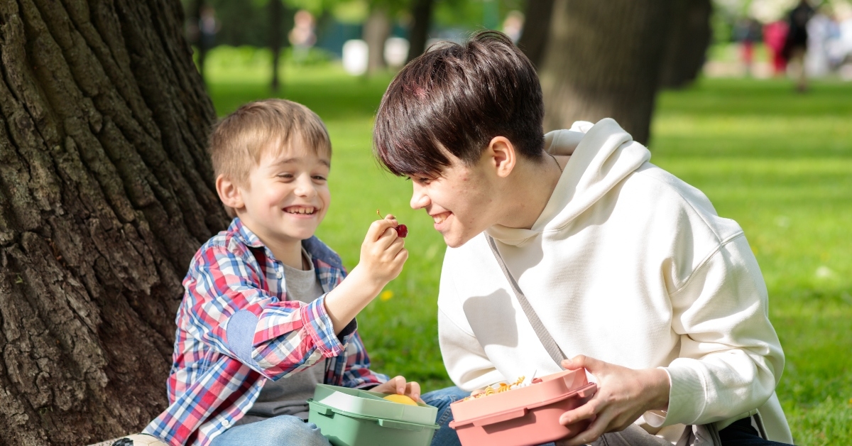 Smaller kid feeding the older one from his lunch box sitting outdoors
