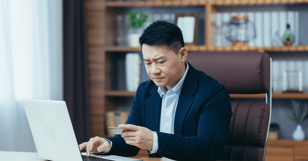 A man stares down at his credit card with his brows furrowed while sitting in his home office.