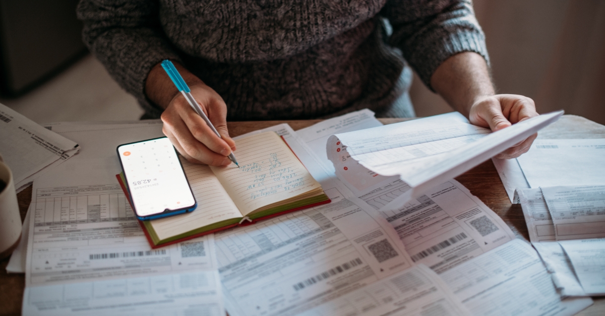 person writing in notebook with calculator and lot of utility bills
