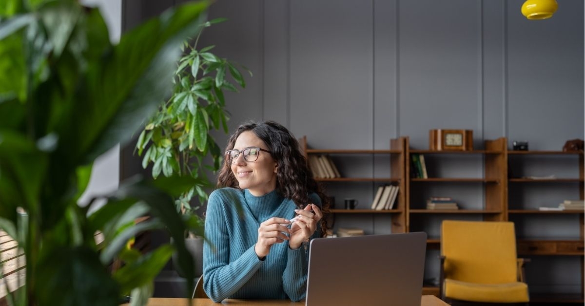 Happy female employee looking in window with satisfied face expression while sitting in office in front of laptop