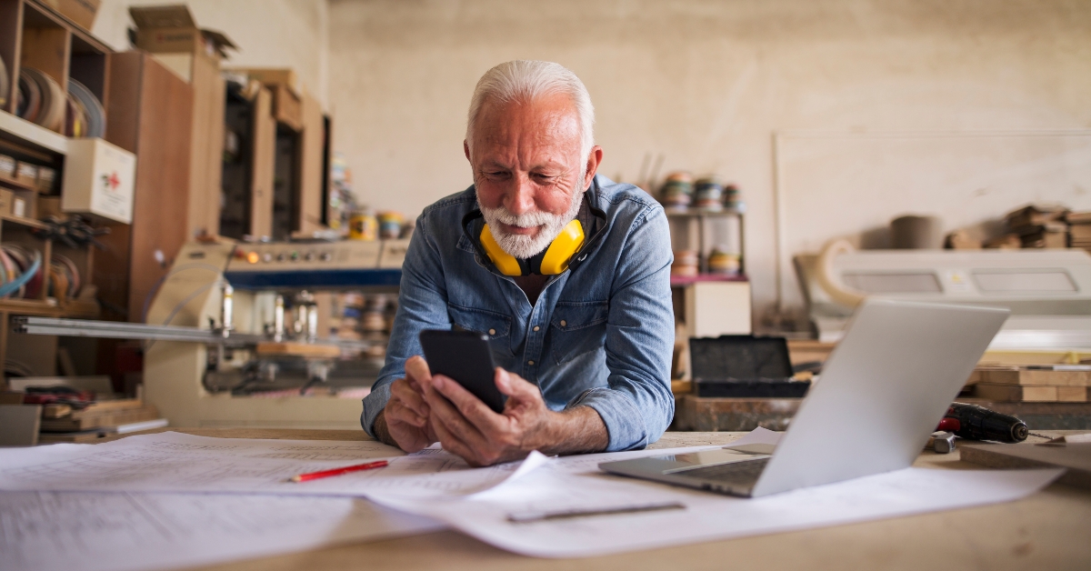 Likeable older carpenter is looking at the cellphone and smiling