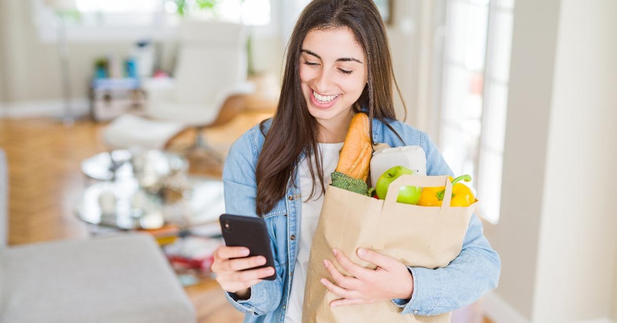 Young woman holding a paper bag full of fresh groceries and using smartphone app for supermarket delivery 