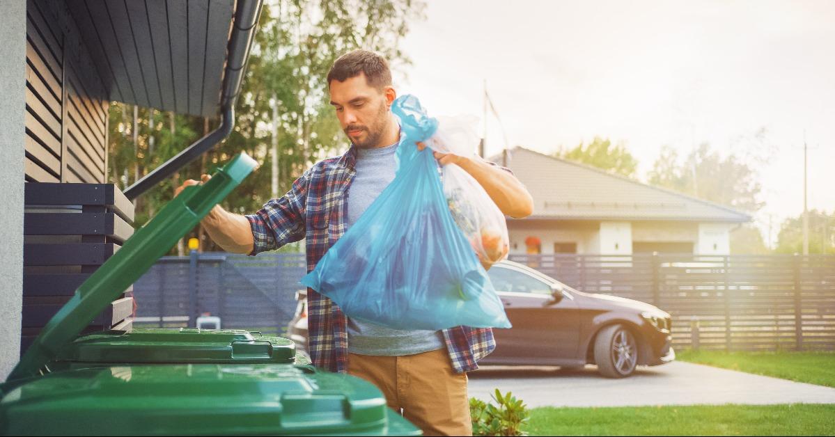 man is throwing away two plastic bags of trash next to his house