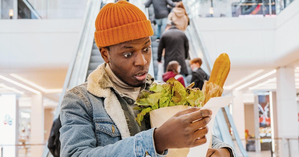 african man with a paper bag of groceries looks upset at a receipt from a supermarket