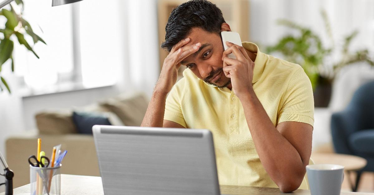 stressed indian man with laptop computer calling on smartphone