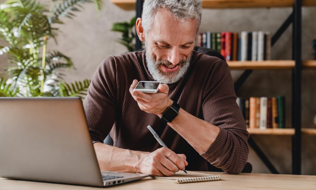man writing notes in notepad while talking by speakerphone