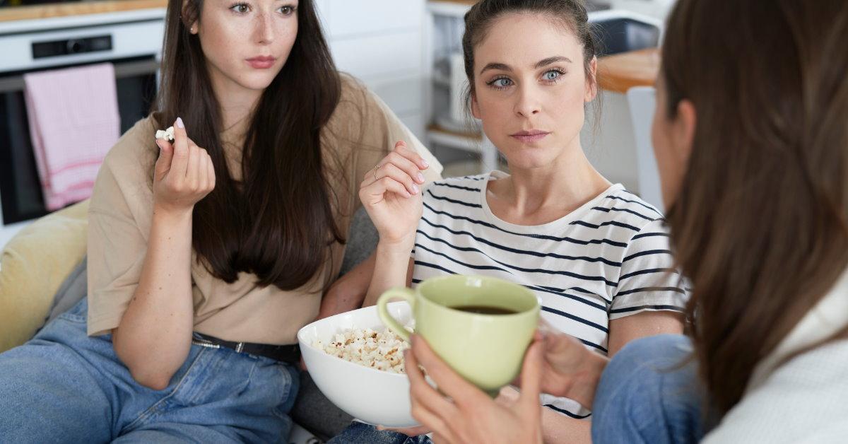 group of three friends sitting on couch talking to each other while eating popcorn