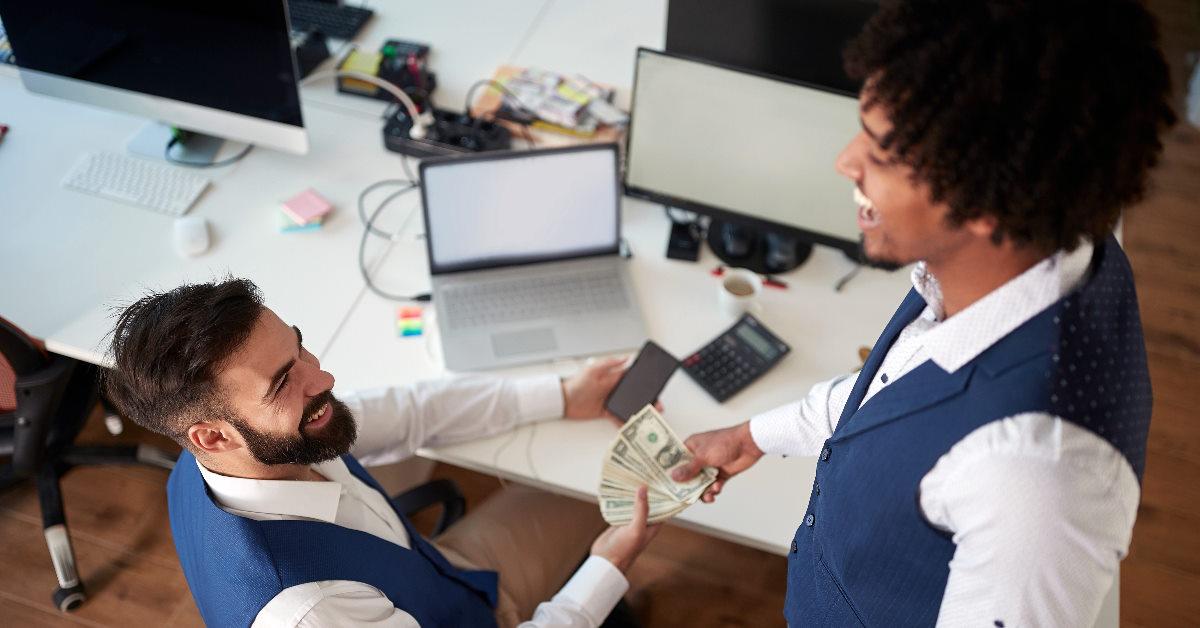 two happy multi ethnic colleagues exchanging money with each other 