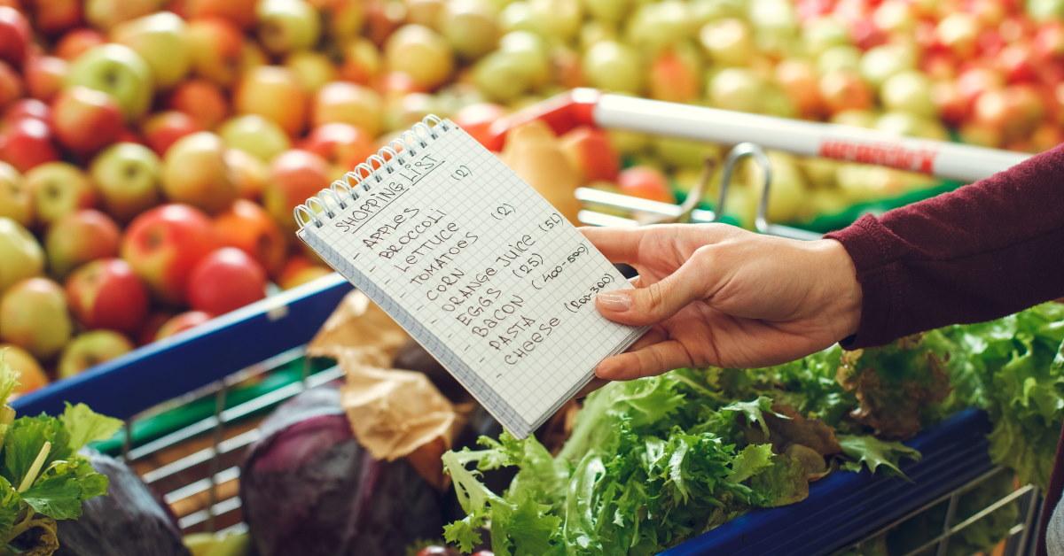 woman holding shopping list besides vegetables cart inside supermarket