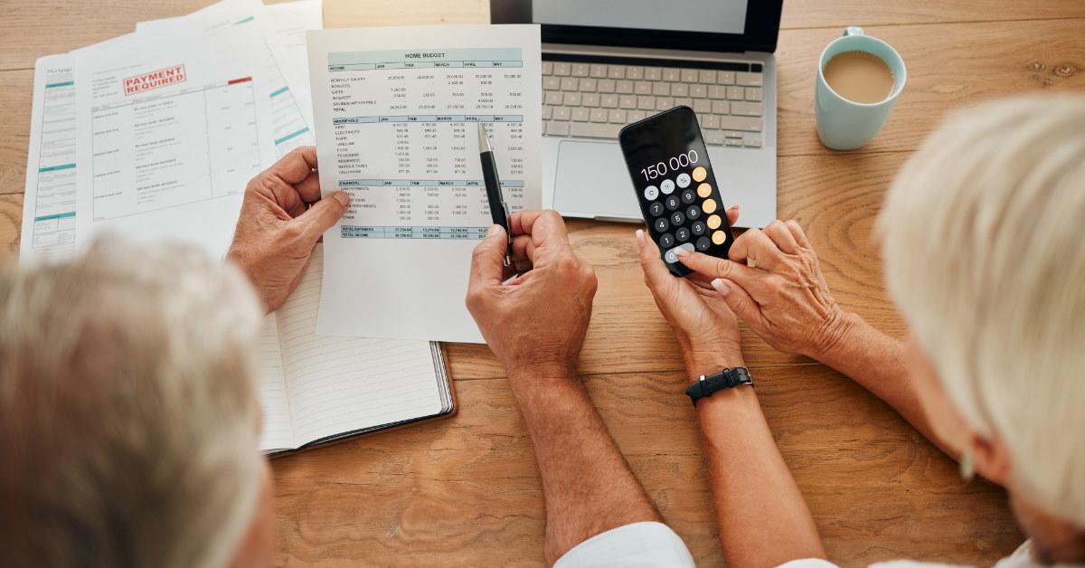 senior couple reviewing bills at table while using calculator on smartphone with laptop 