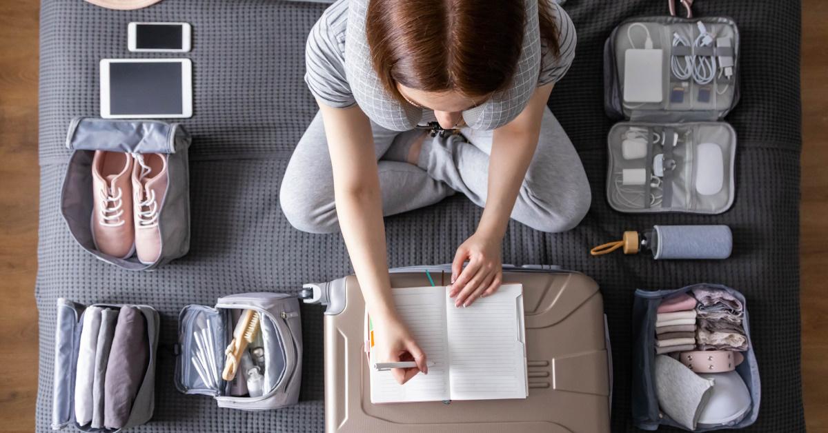 woman sitting on bed using pen and notebook to plan vacation with essentials organized in bags