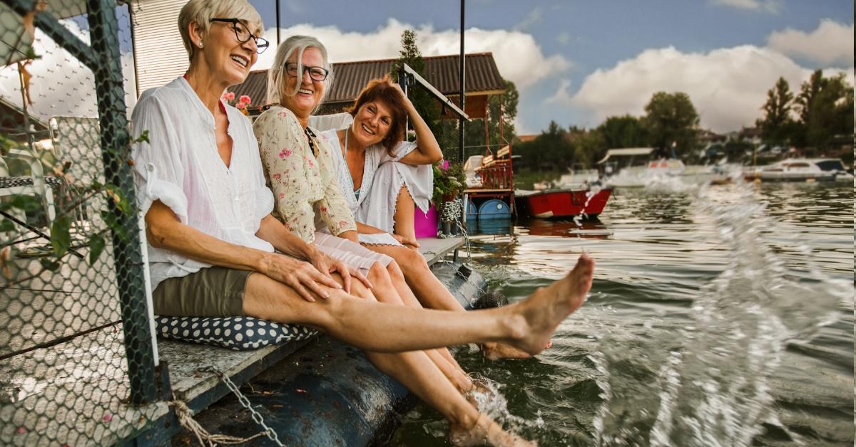senior female friends enjoying vacation while dipping feet in river at cottage