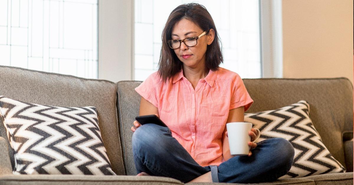 A Mature aged Asian woman using her mobile with coffee in hand and sitting with her legs crossed under her on a sofa