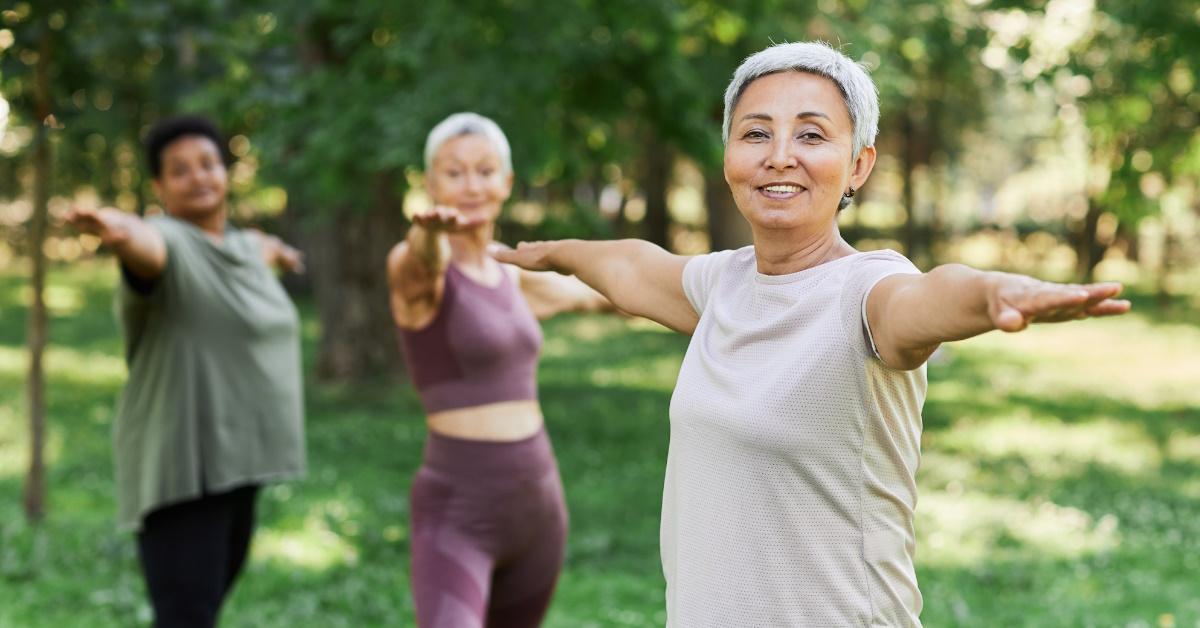 A senior woman is smiling and enjoying a sports workout outdoors with her friends while doing a stretching exercise and looking at the camera.