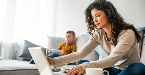 A mom works from her laptop while sitting on the couch next to a young child. 