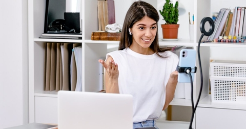 A content creator sits at a desk facing a phone held on a tripod. She is making a video to post on social media. 