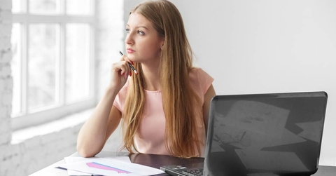 A young adult woman sits and thinks at a desk with a laptop.