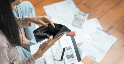 A woman opens an empty black wallet, while bills and paperwork scatter the floor.