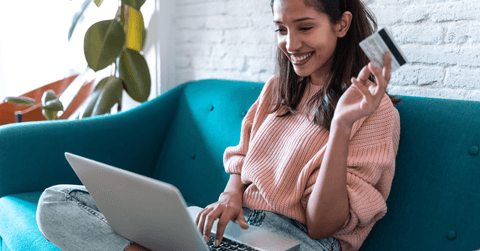 A woman sits on the couch with a laptop in her lap and holding a credit card.