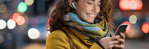 A woman wears wireless earbuds and a scarf while smiling and staring down at her phone.