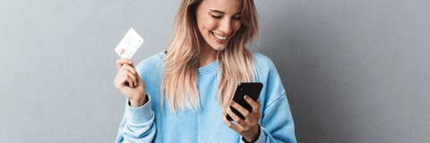 A woman in a blue sweater smiles while holding her phone and credit card. 