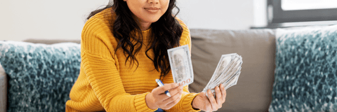 A woman in a yellow sweater sits on the couch while counting a stack of 100 dollar bills.