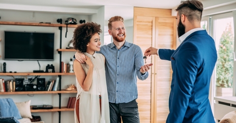 young male real estate agent standing standing inside house handing keys to couple