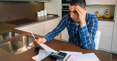 man sitting in home kitchen holding head while stressing about bills at countertop