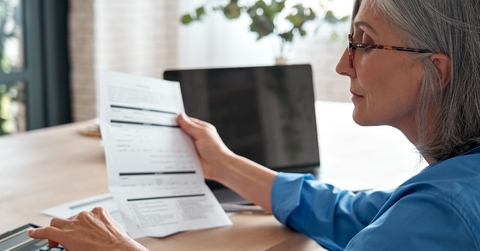 senior lady holding paper bill and calculating the monthly expenses on the calculater while sitting at the table at home.