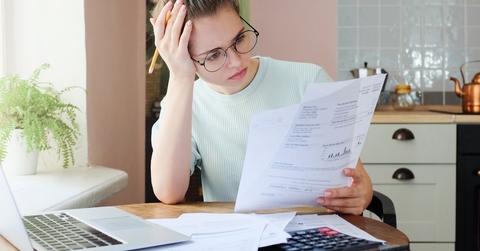  A young girl of European Caucasian descent, with her hair in a bun, is using a calculator to perform calculations while sitting in her home office with a laptop and holding a paper with business data