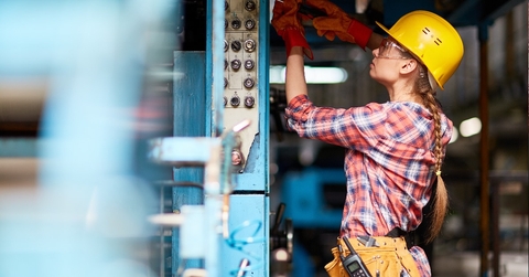 Female technician wearing yellow safety halmet is fixing a part of machinery with screwdriver in factory.