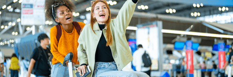 A woman rides atop a luggage cart in the airport while her friend pushes it from behind.