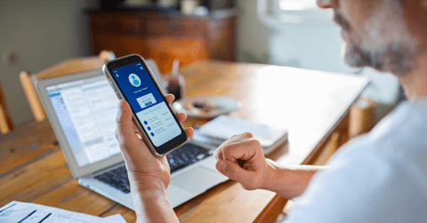 Man checking online bank account balance on smartphone while working on laptop at home