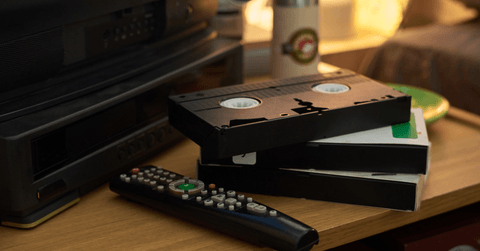 Close-up of several vintage VHS tapes stacked near remote control on wooden table with an old TV