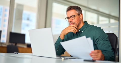 Man in his 40's looking at paperwork and computer thinking